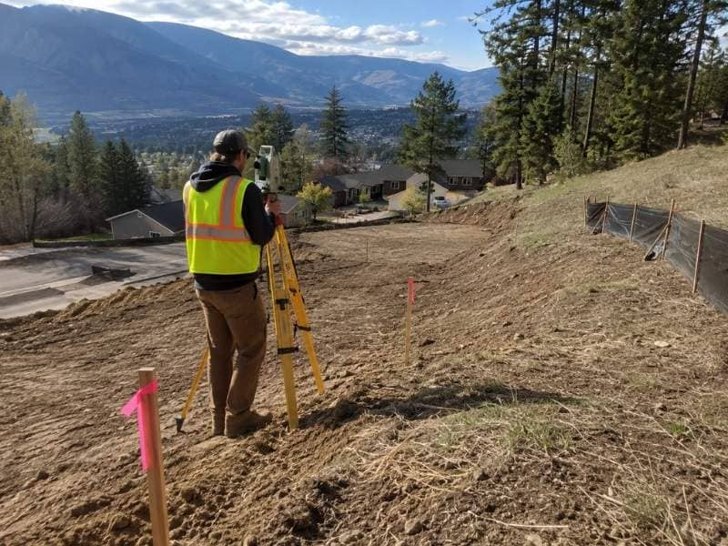 Land surveyor measuring the grade of a steep residential building site near mountain terrain.